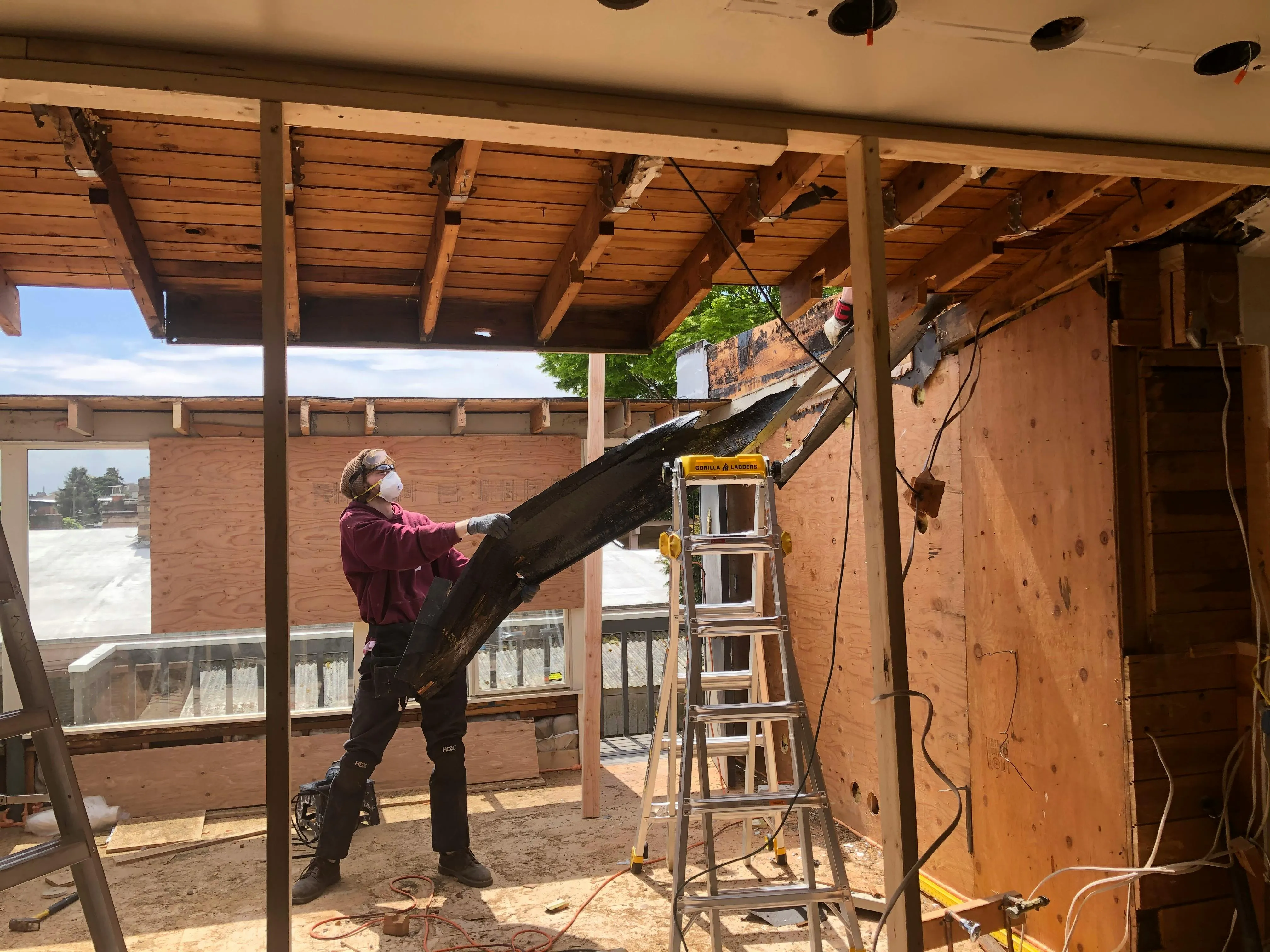 A worker in a respirator mid-demolition, carrying a charred beam out of a torn-open second-story room with sky visible through the framing.