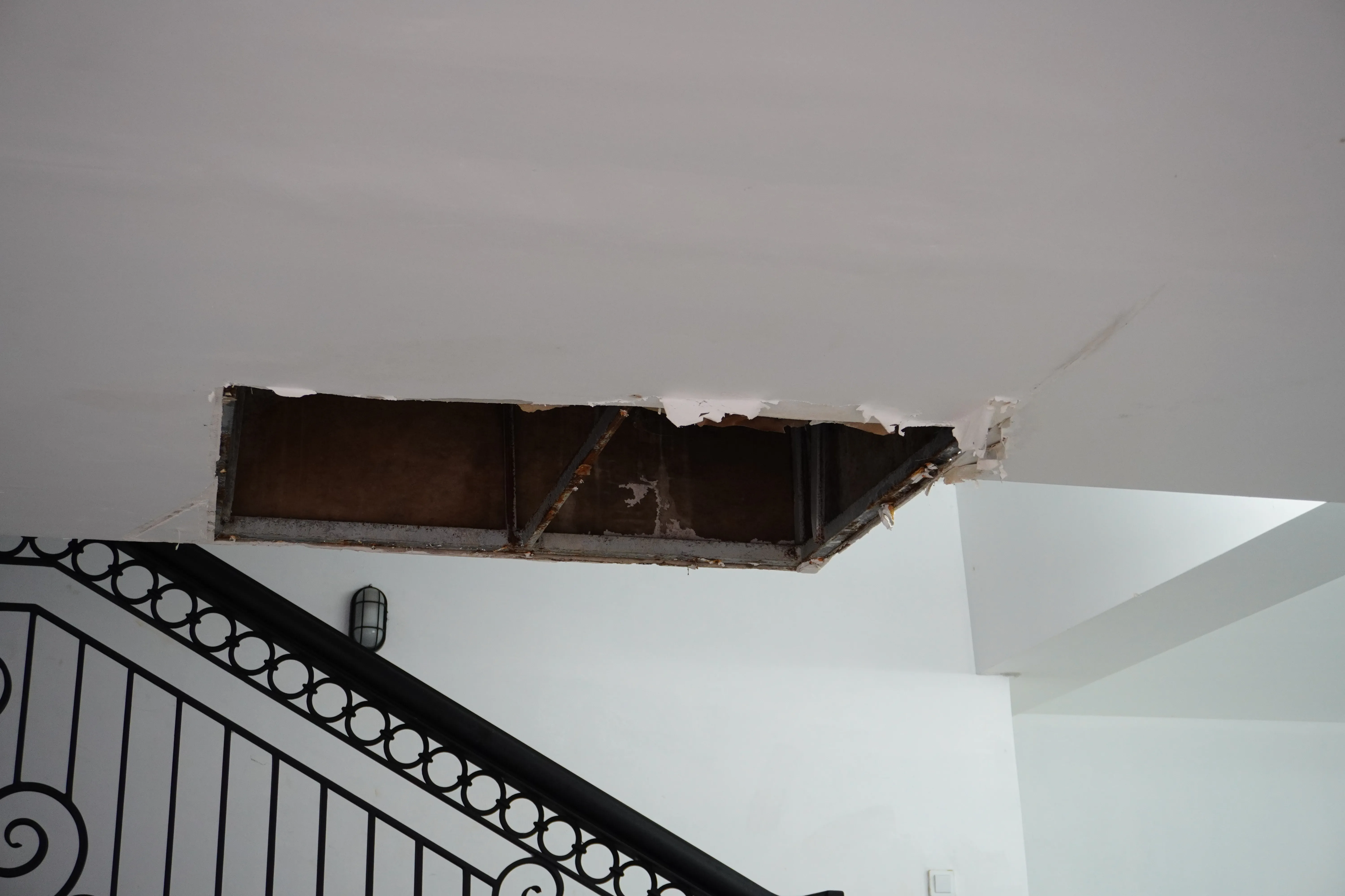 Ceiling cut open above a stairwell to investigate structural framing — torn drywall around the opening exposes dark joist bays and a steel beam underneath, the kind of diagnostic work a structural specialist does before a repair.
